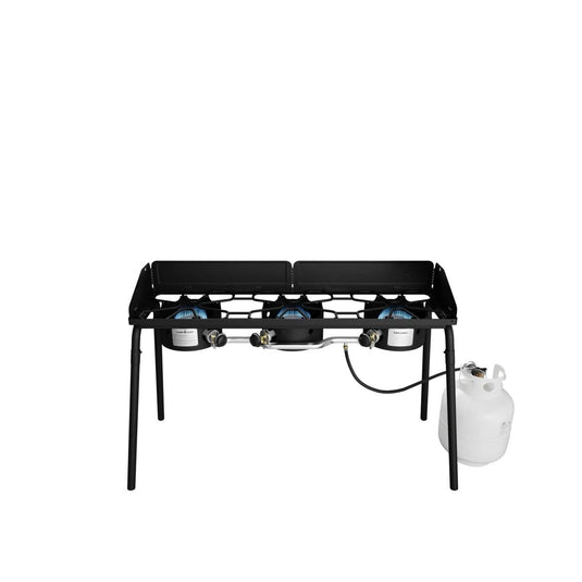 The Camp Chef Explorer is a black, three-burner outdoor stove with four legs and a metal wind guard around the burners. Shown connected to a white propane tank, it offers versatile outdoor cooking on any adventure.