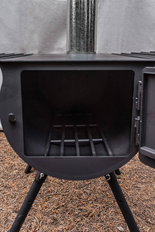 A close-up of the Camp Chef Alpine Heavy Duty Cylinder System, a black metal wood stove with its door open to show the grate inside. Ideal for wall tent camping, it stands on four legs over pine needles with a chimney extending upward.