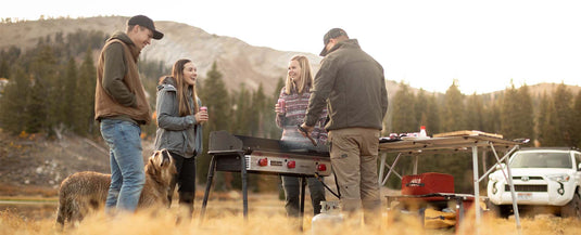 Four people stand outdoors by a grill and folding table, laughing and holding drinks. A dog is nearby, and trees and mountains are in the background. A white vehicle is parked to the right.