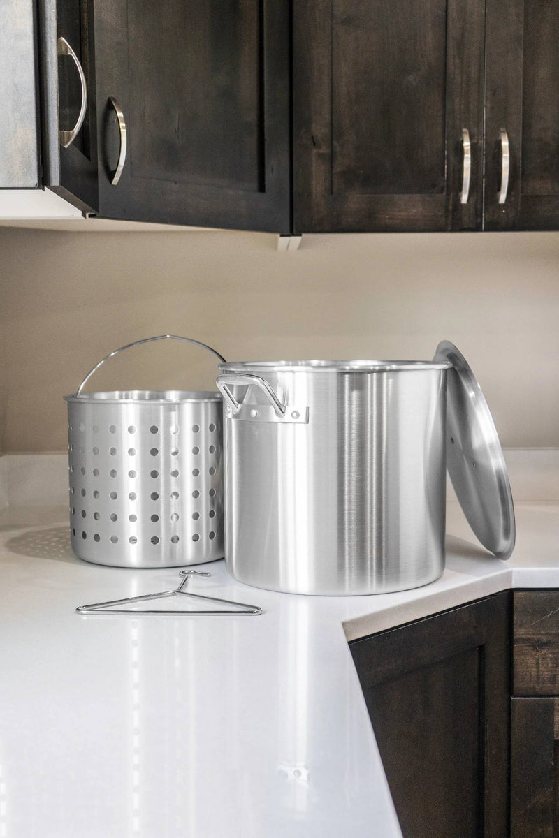 Load image into Gallery viewer, The Camp Chef Aluminum Cooker Pot, featuring a lid, perforated basket, and metal rack, rests on a white kitchen countertop below dark wooden cabinets.
