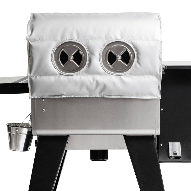 Load image into Gallery viewer, The Camp Chef Woodwind Pro Blanket is a stainless steel grill with a silver fire-resistant fiberglass cover, two circular vents, black legs, and a small metal bucket hanging from the side.
