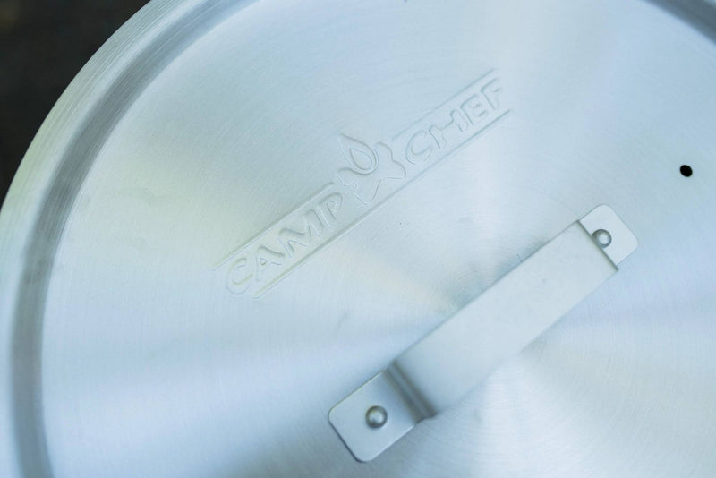 Load image into Gallery viewer, A close-up of the Camp Chef logo embossed on a metal lid with a handle, resting on the Aluminum Cooker Pot by Camp Chef, with a blurred background.
