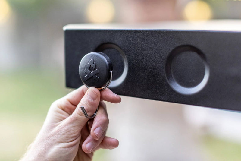 Load image into Gallery viewer, A hand attaches a round black Camp Chef Magnetic Tool Holder from the 3 Pack to a metal surface with two circular indents. The blurred background highlights this branded grilling accessory for organizing tools.
