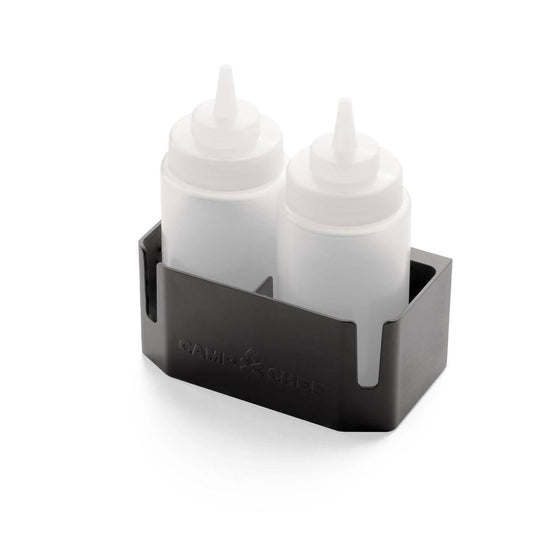 The Camp Chef Bottle Caddy holds two translucent squeeze bottles with pointed caps side by side. It features side slots and has the Camp Chef logo embossed on the front, offering convenient storage for your bottles.