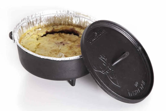 A Camp Chef Disposable Dutch Oven Liner lines a round foil baking dish filled with cobbler, nestled in a black deep Dutch oven with its lid partially open—making Dutch oven cooking easy and mess-free.