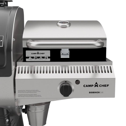 A close-up of a Camp Chef Sidekick Sear attached to a Camp Chef Woodwind 24 grill, highlighting the stainless steel lid with temperature gauge, control knob, and the Camp Chef logo on the front panel.