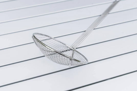 The Camp Chef Long Handled Skimmer, featuring a nickel-plated handle and round mesh basket, rests diagonally on a white slatted surface.