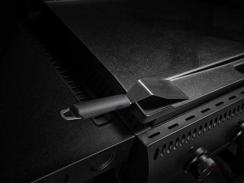 Load image into Gallery viewer, A close-up of the Camp Chef Gridiron 36 shows its flat metal griddle and control knobs. A black spatula rests on the edge, while dim lighting highlights its sleek design and even heating for perfect results every time.
