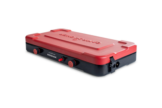The Camp Chef Kodiak is a compact red and black portable camp stove with two burner knobs, offering reliable outdoor performance. Shown on a white background.