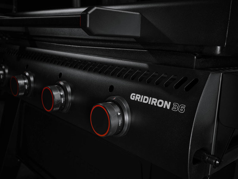 Load image into Gallery viewer, Close-up of the Camp Chef Gridiron 36 grill, highlighting four black control knobs with red accents. Dramatic lighting emphasizes its sleek design, spacious griddle for even heating, and easy clean-up. "Gridiron 36" is visible on the front panel.
