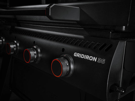 Close-up of the Camp Chef Gridiron 36 grill, highlighting four black control knobs with red accents. Dramatic lighting emphasizes its sleek design, spacious griddle for even heating, and easy clean-up. "Gridiron 36" is visible on the front panel.
