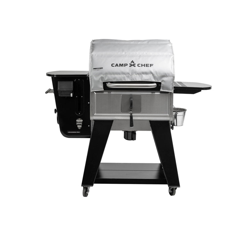 Load image into Gallery viewer, A Camp Chef Woodwind Pro Blanket grill, featuring a silver cover, black side shelf, lower storage shelf, wheels, and a fire-resistant fiberglass blanket, is displayed against a white background.
