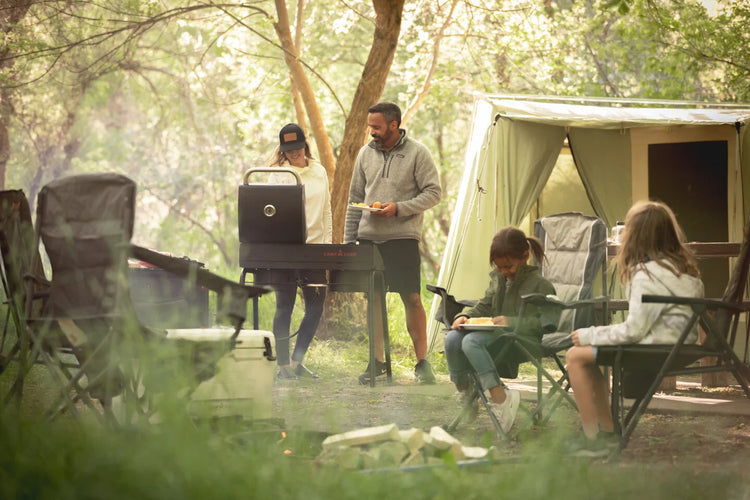 Four people are camping in a forest. Two stand by a grill, preparing food, while two sit in chairs eating. A large tent is set up behind them, surrounded by trees and camping gear in a sunlit, green setting.