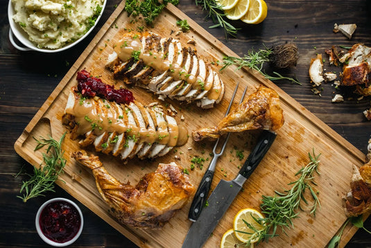 Sliced roast turkey with gravy and cranberry sauce on a wooden cutting board, surrounded by turkey legs, lemon slices, rosemary, mashed potatoes, and cranberry sauce. Two knives rest beside the turkey.