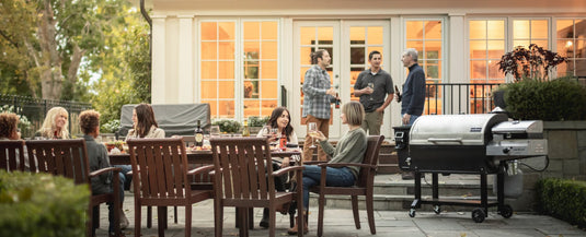 A group of people enjoy a backyard barbecue, with some sitting at a wooden outdoor table eating and talking, while others stand and chat near a grill on a patio in front of a well-lit house.