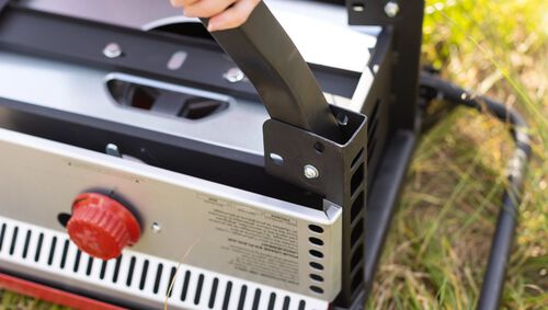 Close-up of a portable grill with a hand holding its handle, set against a natural background.