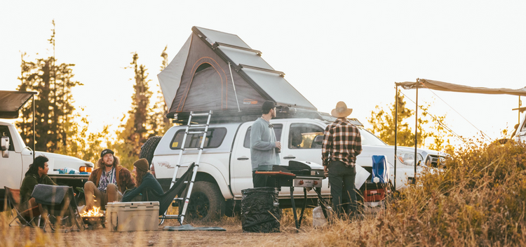 A group of people are camping outdoors near a white truck with a rooftop tent. Some sit by a campfire, while others stand and cook at a grill. Trees and camping gear surround them under a clear sky.