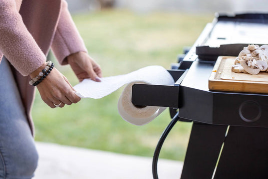 A person pulls a sheet from the Camp Chef Magnetic Paper Towel Holder attached to an outdoor grill, while a cutting board with sliced mushrooms sits on the side shelf, showcasing efficient workspace organization.