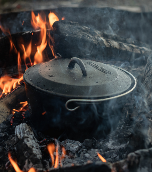 A black cast iron pot with a lid sits over an open campfire, surrounded by burning wood and smoke.