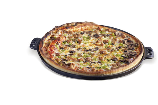 A whole baked pizza with cheese, mushrooms, and green peppers sits on a Camp Chef Cast Iron Pizza Pan with decorative handles, isolated on a white background.