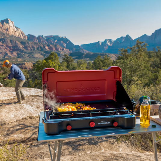 Person cooking outdoors on a camping stove with scenic mountains in the background