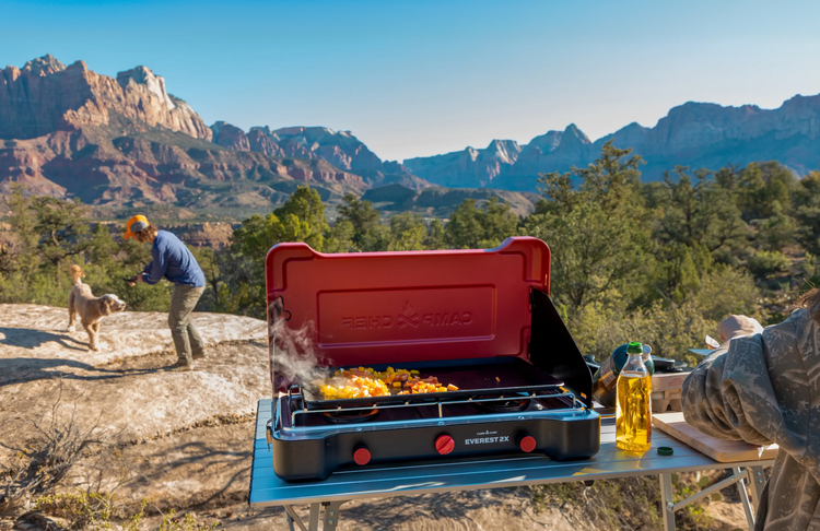 Person cooking outdoors on a camping stove with scenic mountains in the background