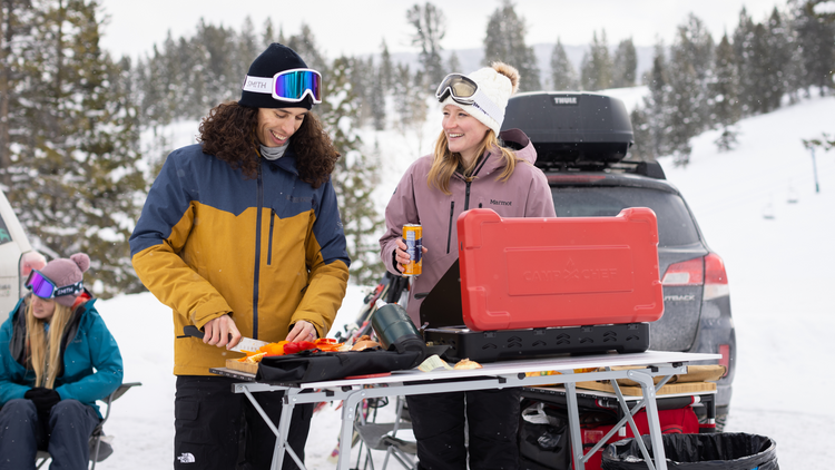 Two people wearing winter jackets and goggles smile while preparing food at an outdoor table with a camping stove. Snowy trees and a parked car are in the background; two others sit together nearby.