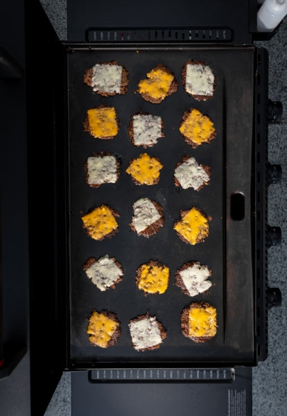 A top-down view of a griddle with 18 burger patties, each topped with either white or yellow melted cheese, arranged in rows and cooking on the flat surface.