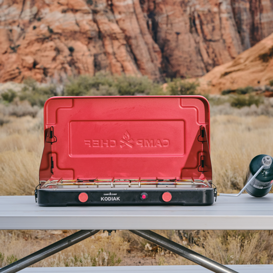 A red Camp Chef Kodiak portable camp stove sits on a metal table outdoors, with desert vegetation and large red rock formations in the background. A small propane tank is attached on the right side.