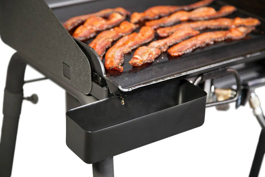 Close-up of sizzling bacon strips cooking on a griddle, with grease draining efficiently into the Camp Chef Professional Grease Cup below for optimal management.