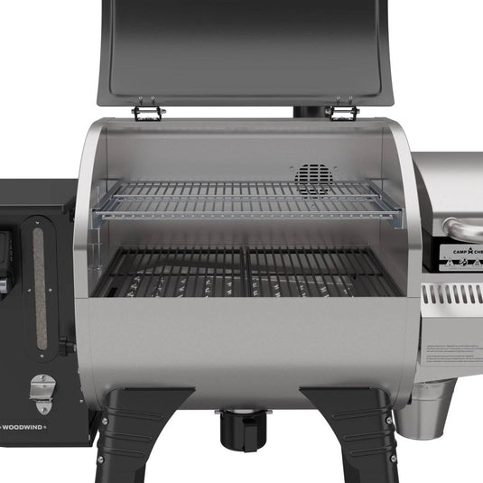 A close-up of the Camp Chef Woodwind 24 pellet grill with its lid open, displaying black grates on the main area and a raised second rack. This stainless steel grill features sturdy legs, metal parts, and a WIFI controller for easy use.