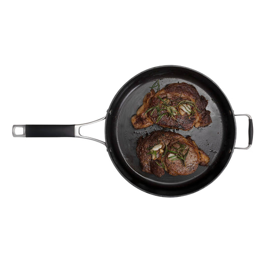 A top-down view of the Camp Chef Heritage Skillets, a black cast iron frying pan with black and metal handles, featuring two cooked steaks garnished with rosemary and garlic, renowned for excellent searing, set against a white background.