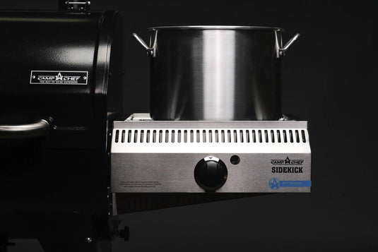A large stainless steel pot sits on the propane burner of a black Camp Chef Sidekicks, a versatile grill accessory, against a dark background.