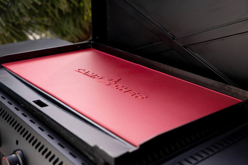 Load image into Gallery viewer, A close-up of a red Camp Chef grill lid with the brand's flame logo, featuring a Silicone Griddle Mat for added protection. The grill is outdoors with blurred trees in the background.
