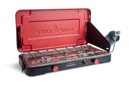 The Camp Chef Kodiak portable two-burner propane stove features a red lid, black base, and attached propane tank on the right. The open lid reveals cooking grates and control knobs on this 20,000 BTU camp stove.