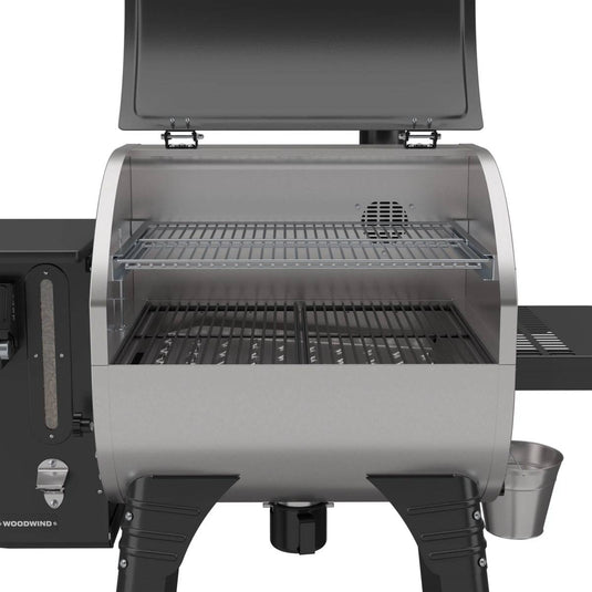 The Camp Chef Woodwind 24 pellet grill, shown with the lid open and two cooking racks, features a WIFI controller, black legs, and a side shelf with a silver bucket—making outdoor grilling easy and convenient.