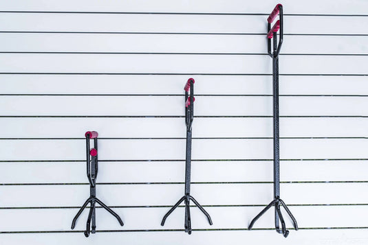 Three Camp Chef Dutch Oven Lid Lifters in varying lengths, with black shafts and red handles/tips, hang vertically on a white slatted wall—ideal for lifting cast iron lids, arranged shortest to tallest.