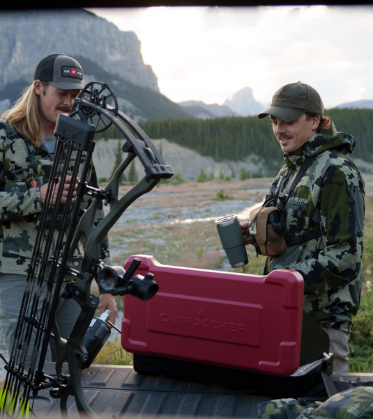 Two men in camo clothing stand by a red Camp Chef cooler on a truck bed, holding mugs and a bow, with mountains, trees, and a river in the background.