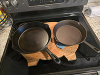 Two cast iron skillets sit side by side on a stovetop, resting on a wooden cutting board shaped like a pig. The stove is black with silver knobs, and a butter dish and spice rack are visible on the granite countertop.