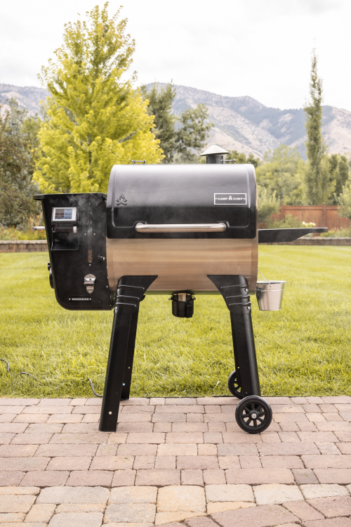 A black and stainless steel pellet grill smoker stands on a patio with green grass, trees, and mountains in the background. Smoke is coming from the grill’s chimney, and a small metal bucket hangs from the side.