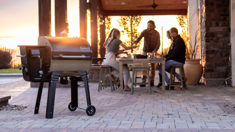 A grill stands on a patio in the foreground, while three people sit at an outdoor table enjoying a meal together at sunset, surrounded by a relaxed, warm atmosphere.