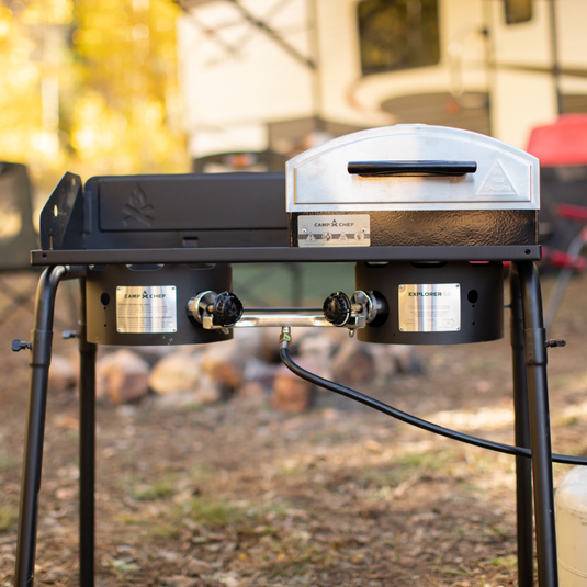 A portable outdoor gas grill and pizza oven setup stands on grass at a campsite, with a blurred RV and trees in the background. The grill features dual burners and visible control knobs.