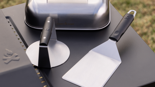 A metal pizza cutter and a flat spatula with black handles rest on a dark outdoor grill surface, with a metallic container and green grass visible in the background.